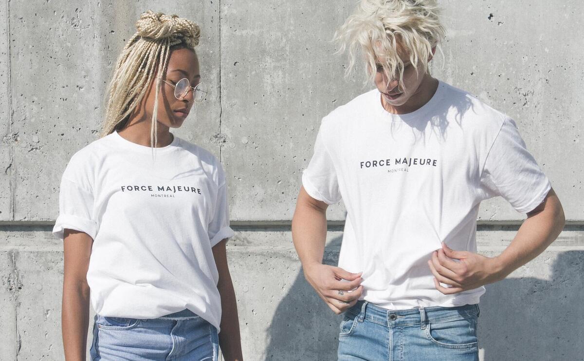 man and woman in white-and-gray force majeure-printed crew-neck t-shirts standing near gray concrete wall