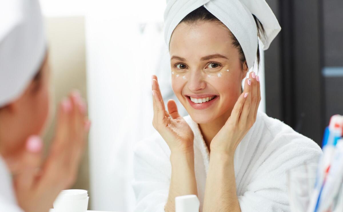 Happy woman applying moisturizer around eye in bathroom.