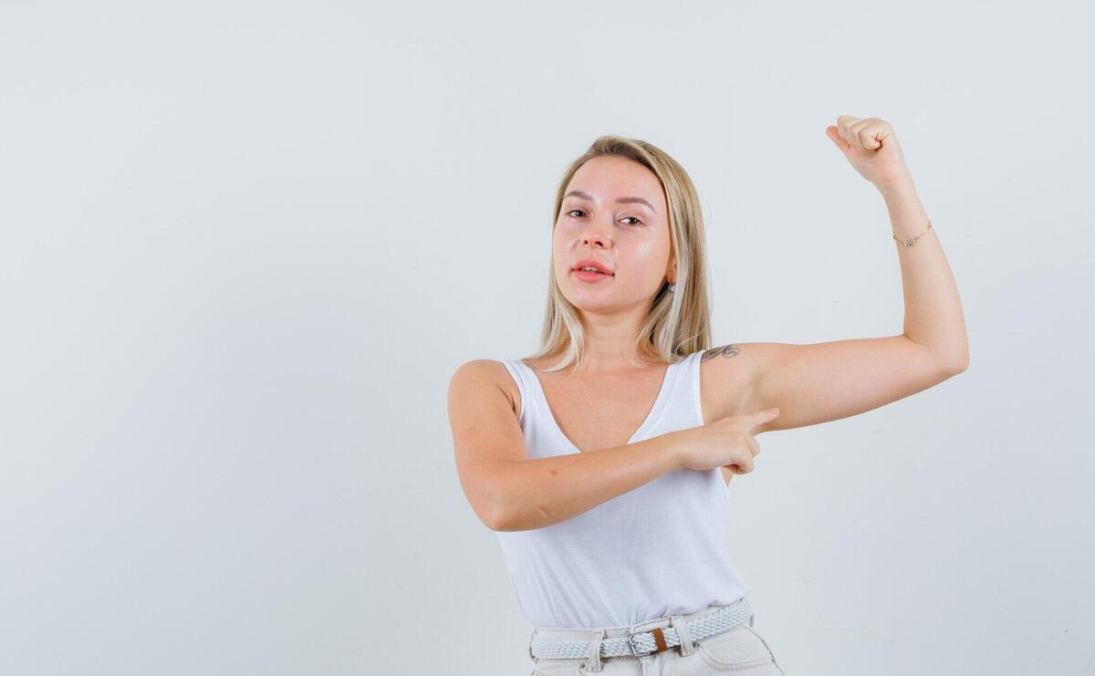 Young lady in white blouse pointing at her arm muscles and looking focused